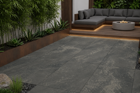Large rectangular outdoor patio pavers in a dark gray stone-look finish, installed in a modern backyard patio setting with a gray sectional sofa, wooden deck, and greenery.